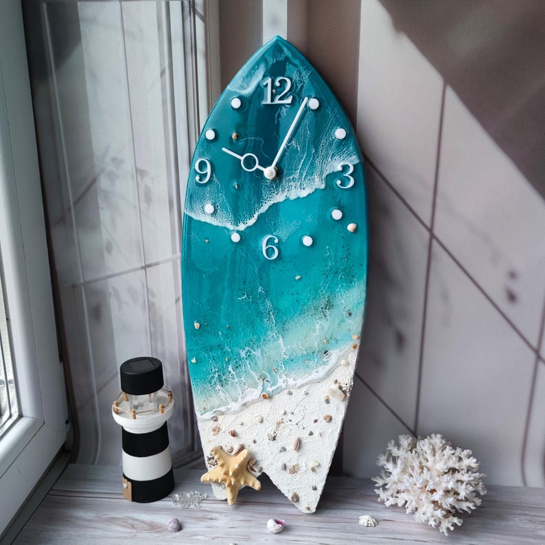 Peut inclure: Horloge murale en forme de planche de surf avec un design sur le th&egrave;me de la plage. Le cadran de l'horloge pr&eacute;sente une sc&egrave;ne d'oc&eacute;an turquoise avec des vagues blanches, des coquillages et une &eacute;toile de mer. Des chiffres blancs indiquent les heures.