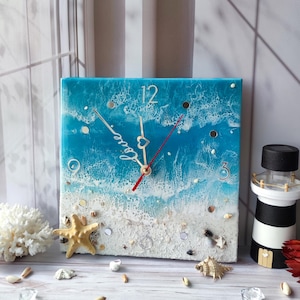 May include: Square clock with a blue and white ocean wave design. The clock face has white numbers and a red and silver hand. The clock is surrounded by white sand and seashells.