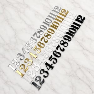 Può includere: Una collezione di numeri metallici in argento, oro, bianco e nero, disposti diagonalmente su una superficie di marmo. I numeri, da 1 a 12, sono adatti a vari progetti di artigianato e design.