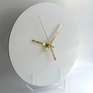May include: A modern, minimalist white wall clock with gold-colored hands and a black rim. The clock is displayed on a clear acrylic stand, showcasing its clean design. The clock's face is a simple, uncluttered white.