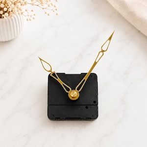 May include: A black square clock movement mechanism featuring two gold-coloured metal clock hands. The hands have an open, elongated diamond-shaped design at the tips and are attached to the centre of the black plastic housing unit.
