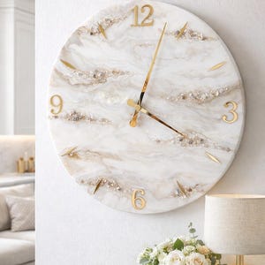May include: A round wall clock with a white marble-like face, gold numerals, and gold hands. The clock features embedded gold and crystal accents. The clock is mounted on a white wall, with a lamp and flowers in the foreground.