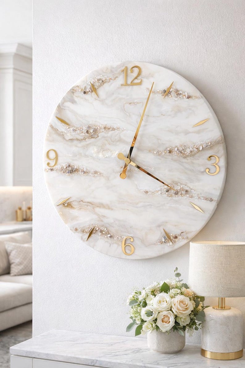 May include: A round wall clock with a white marble-like face, gold numerals, and gold hands. The clock features embedded gold and crystal accents. The clock is mounted on a white wall, with a lamp and flowers in the foreground.