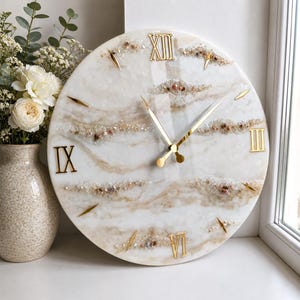 May include: A round wall clock with a white and beige marbled resin finish. The clock face features gold-coloured Roman numerals at the twelve, three, six, and nine o'clock positions, gold hands, and horizontal bands of embedded crushed crystals.