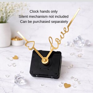 Puede incluir: Agujas de reloj doradas, una con forma de coraz&oacute;n, y la palabra "love" en cursiva. Las agujas est&aacute;n unidas a un mecanismo de reloj negro. El texto de la imagen dice: "Solo agujas. Mecanismo silencioso no incluido. Se puede comprar por separado."