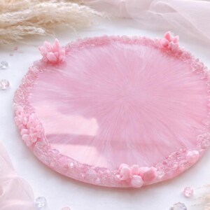 May include: A round, pink resin tray with a textured surface. The tray is decorated with small, pink crystal-like embellishments around the edge and three floral accents. The tray is a light pink color and has a glossy finish.