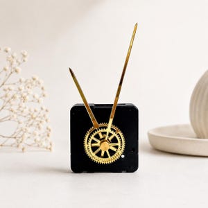 May include: A black square clock movement mechanism featuring a gold-coloured gear-shaped centre piece and two long, slender, pointed gold-coloured clock hands extending upwards in a V-shape from the central gear assembly.