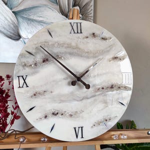 May include: Round wall clock with a white and gray marbled design. The clock face features black Roman numerals at the IX, XII, and VI positions, with silver-colored hands. Decorative crystals and accents are embedded in the design.