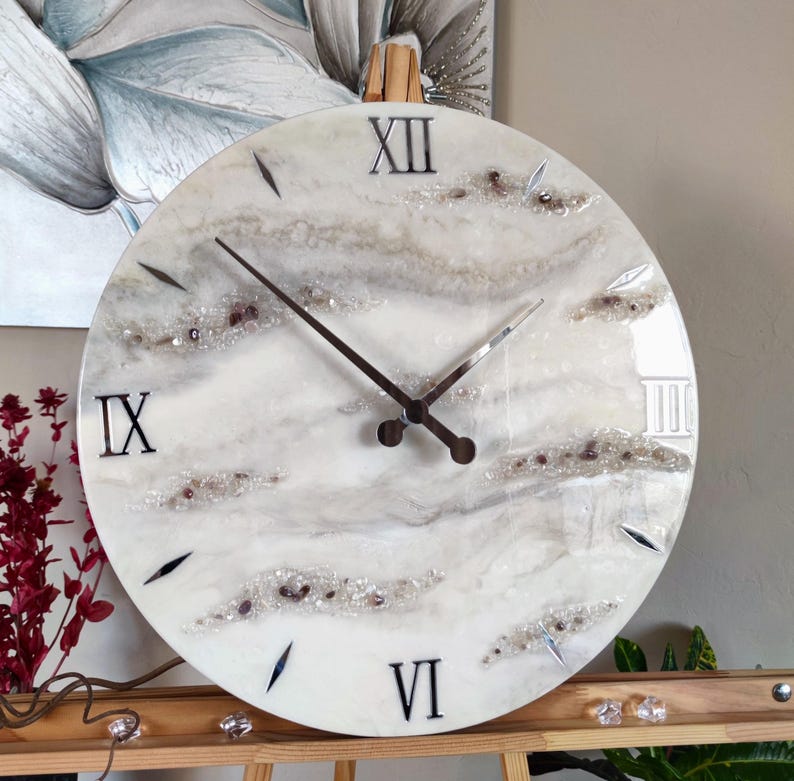 May include: Round wall clock with a white and gray marbled design. The clock face features black Roman numerals at the IX, XII, and VI positions, with silver-colored hands. Decorative crystals and accents are embedded in the design.