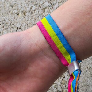 Pode incluir: Uma pulseira com listras azuis, amarelas e rosa. A pulseira é feita de tecido e tem um fecho de metal.