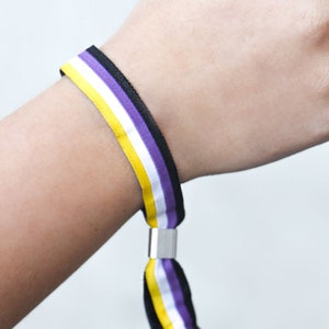 Puede incluir: Una pulsera de rayas moradas, blancas y amarillas con ribete negro. La pulsera tiene un cierre de metal.