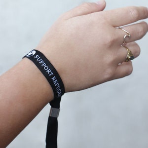 Pode incluir: Uma pulseira preta com texto branco que diz "SUPPORT REFUGEES".