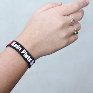 Może przedstawiać: Czarna opaska na nadgarstek z białym napisem "Kein Platz für", niemieckie wyrażenie oznaczające "Brak miejsca dla".