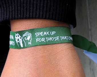 May include: A green wristband with the text "Speak Up For Those That Can't" and a raised fist graphic. The wristband is worn on a person's wrist.