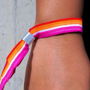 May include: A pink, white, orange and red striped wristband with a silver clasp. The wristband is worn on a person's wrist.