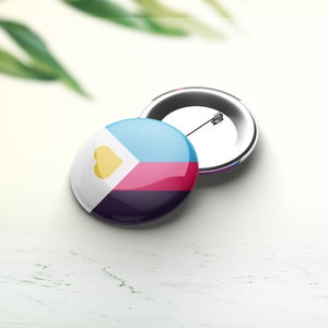 Op de afbeelding: Twee ronde buttons. Eén button toont een ontwerp met lichtblauwe, witte, roze en donkerblauwe secties en een gele hart. De andere button toont de speld en de achterkant.