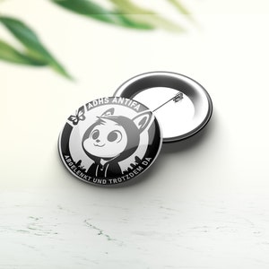 Peut inclure: Badge bouton noir et blanc avec un personnage de dessin animé portant un sweat à capuche et le texte "ADHS ANTIFA ABGELENKT UND TROTZDEM DA".