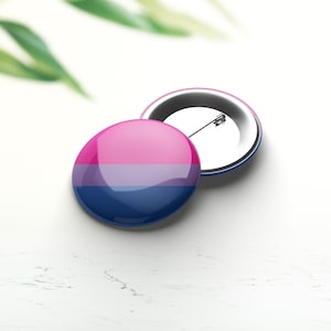 Può includere: Una spilla da bottone con i colori della bandiera bi, con strisce orizzontali rosa, azzurro e blu scuro. La spilla è su una superficie bianca.