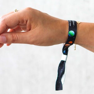 Könnte beinhalten: Ein schwarz-weißes Armband mit einem bunten Planeten-Design und dem Text "Unser Planet, unser einer." Das Armband ist um das Handgelenk einer Person gebunden.