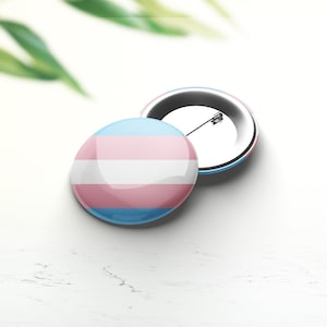 Könnte beinhalten: Eine Transgender-Pride-Flaggennadel mit blauem, weißem und rosafarbenem Design. Die Nadel befindet sich auf einer weißen Oberfläche.