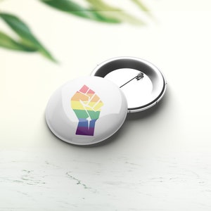 Könnte beinhalten: Ein weißer Knopf mit einer Regenbogenfaust, die in die Luft erhoben ist. Der Knopf ist an einer silbernen Sicherheitsnadel befestigt.