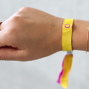 Könnte beinhalten: Ein gelbes Armband mit einem rosa Kreis und ein goldenes Kettenarmband mit einem kleinen runden Anhänger.