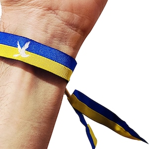 Könnte beinhalten: Ein blaues und gelbes Armband mit einem weißen Taubensymbol. Das Armband ist um das Handgelenk einer Person gebunden.