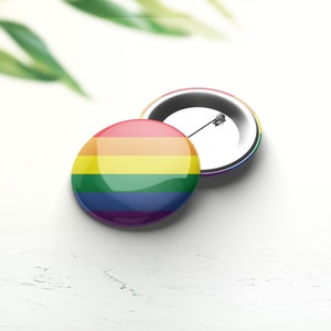 Könnte beinhalten: Zwei runde Ansteckbuttons mit Regenbogenmuster, die die LGBTQ+-Pride-Flagge darstellen. Ein Button ist nach oben, der andere nach unten gerichtet und zeigt die Nadel. Die Buttons befinden sich auf einer weißen Oberfläche.