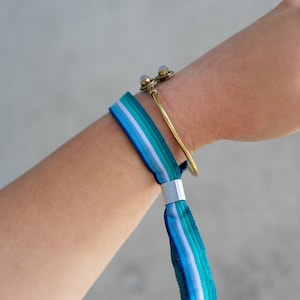 May include: A blue, white and green striped wristband with a silver clasp. The wristband is worn on a person's wrist with a gold bracelet.