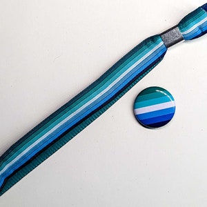 Op de afbeelding: Een blauw, turquoise en wit gestreepte polsband met een zilveren metalen sluiting en een bijpassende ronde knoop. De knoop en de polsband hebben een gradiëntontwerp met verschillende tinten blauw en turquoise.