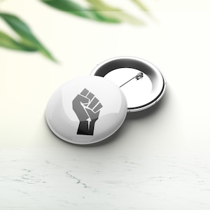 Peut inclure: Un bouton blanc avec un symbole de poing levé noir. Le bouton est attaché à un dos d'épingle argenté.