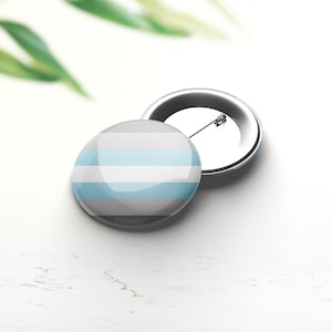 Könnte beinhalten: Zwei runde Ansteckbuttons. Ein Button ist nach oben, der andere nach unten gerichtet. Die Buttons haben ein Design aus horizontalen Streifen in Hellblau, Weiß und Grau. Die Anstecknadeln sind silberfarben. Der Hintergrund ist weiß mit grünen Blättern.