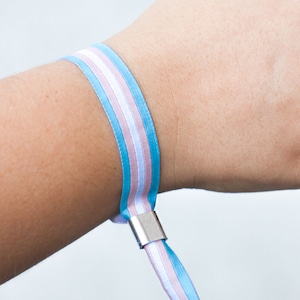 Op de afbeelding: Een blauw, wit en roze gestreepte polsband met een zilveren sluiting. De polsband is om een pols gewikkeld. De kleuren vertegenwoordigen de transgender pride vlag.