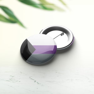 Peut inclure: Un bouton rond avec un design noir, gris et violet. Le bouton représente le drapeau de la fierté asexuelle.