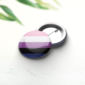 Pode incluir: Um botão redondo com as cores da bandeira do orgulho genderqueer. O botão é rosa, branco, lavanda, preto e azul. O botão está sobre uma superfície branca.