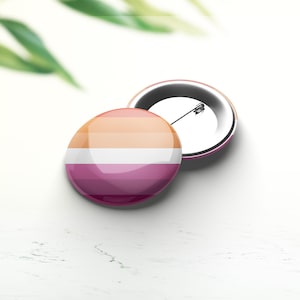 Può includere: Un bottone rotondo con un design di bandiera del pride lesbico. Il bottone è arancione, bianco e rosa. Il bottone è su una superficie bianca.
