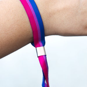 Op de afbeelding: Een armband met een roze, paarse en blauwe lintontwerp. De armband heeft een zilveren metalen sluiting.