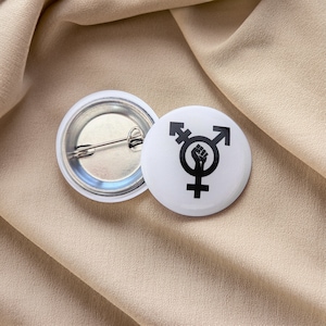 Könnte beinhalten: Ein weißer Knopf mit einem schwarzen Transgender-Symbol und einer erhobenen Faust im Kreis. Der Knopf befindet sich auf einem beigefarbenen Stoffhintergrund.
