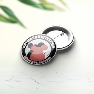 Può includere: Un bottone rotondo bianco con un cerchio nero e una silhouette di topo rossa. Il testo "ANTIFASCHISTISCHE SUDMAUS AKTION" è scritto in bianco attorno al cerchio.