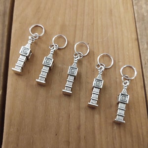 Pode incluir: Cinco chaveiros prateados com pingentes Big Ben. Cada chaveiro apresenta uma réplica em miniatura detalhada da icónica torre do relógio, completa com um pequeno anel para prender chaves. Os chaveiros são exibidos numa superfície de madeira.