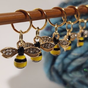 Puede incluir: Cuatro marcadores de punto de abeja dorados con detalles de pedrería transparente. Las abeilles son amarillas y negras con alas doradas. Los marcadores están en una varilla de madera.