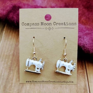 May include: A pair of gold-tone earrings featuring a white enamel sewing machine charm with rhinestones. The earrings are displayed on a brown card with the text "Compass Moon Creations" and a website address.