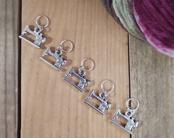 Pode incluir: Cinco marcadores de ponto de costura de máquina de coser prateados em uma superfície de madeira. Os marcadores estão dispostos em uma fila de cima para baixo.
