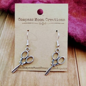 May include: Silver scissor earrings on a brown card with the text "Compass Moon Creations" and a website address.