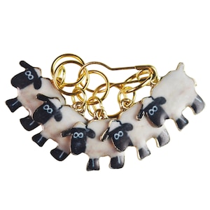 May include: A gold-colored knitting stitch marker set featuring six sheep charms with black faces and white bodies. The sheep are attached to a safety pin and several small rings.