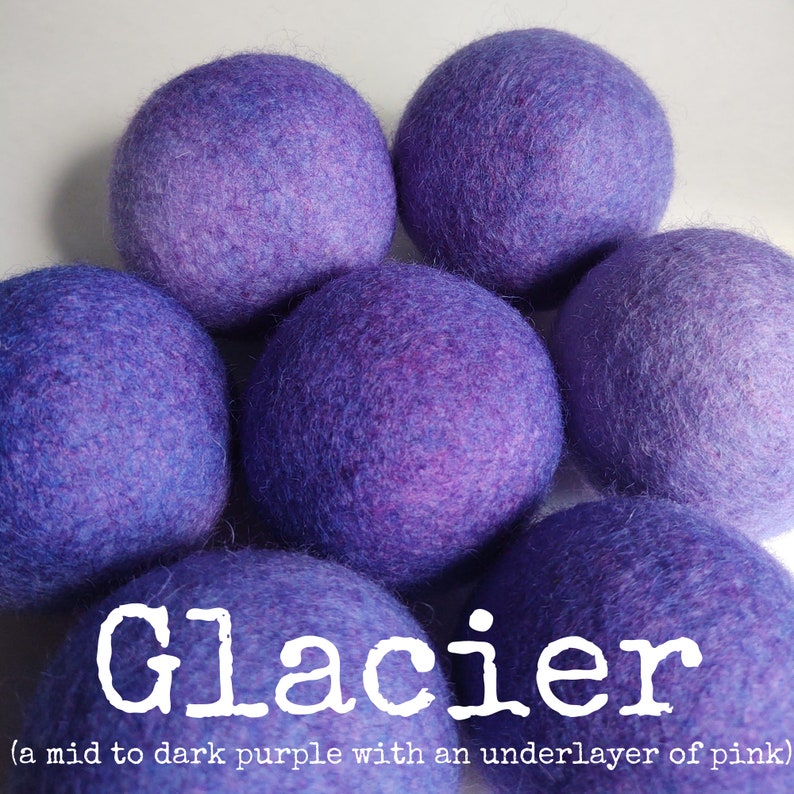 May include: A group of six purple wool dryer balls with a pink undertone. The balls are arranged in a pyramid shape. The text "Glacier (a mid to dark purple with an underlayer of pink)" is below the balls.