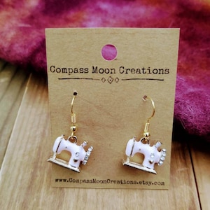 May include: A pair of gold-toned dangle earrings featuring miniature white sewing machines with rhinestone accents. The earrings hang from gold-colored hooks and are displayed on a brown card with the text "Compass Moon Creations".