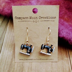 May include: Black enamel sewing machine earrings with gold accents and clear rhinestones. The earrings are on a card with the text "Compass Moon Creations" and a website address.