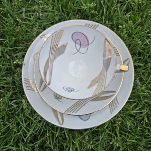 Peut inclure: Un ensemble de tasse et soucoupe en porcelaine blanche avec une bordure dorée et un motif abstrait délicat en or, rose et vert. Le motif présente des lignes et des volutes stylisées.