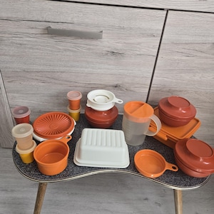 Può includere: Una collezione di articoli da cucina Tupperware vintage nei toni arancione, bianco e plastica trasparente. Include ciotole, contenitori con coperchi, un colino e un misurino. Gli articoli sono disposti su un tavolino.